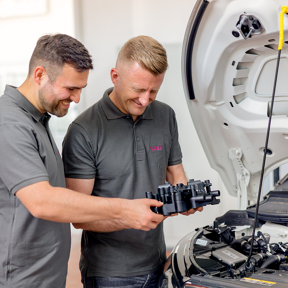 Zwei VOSS Mitarbeiter schauen auf VOSS Modul in ihren Händen, neben einem Pkw mit geöffneter Motorhaube.