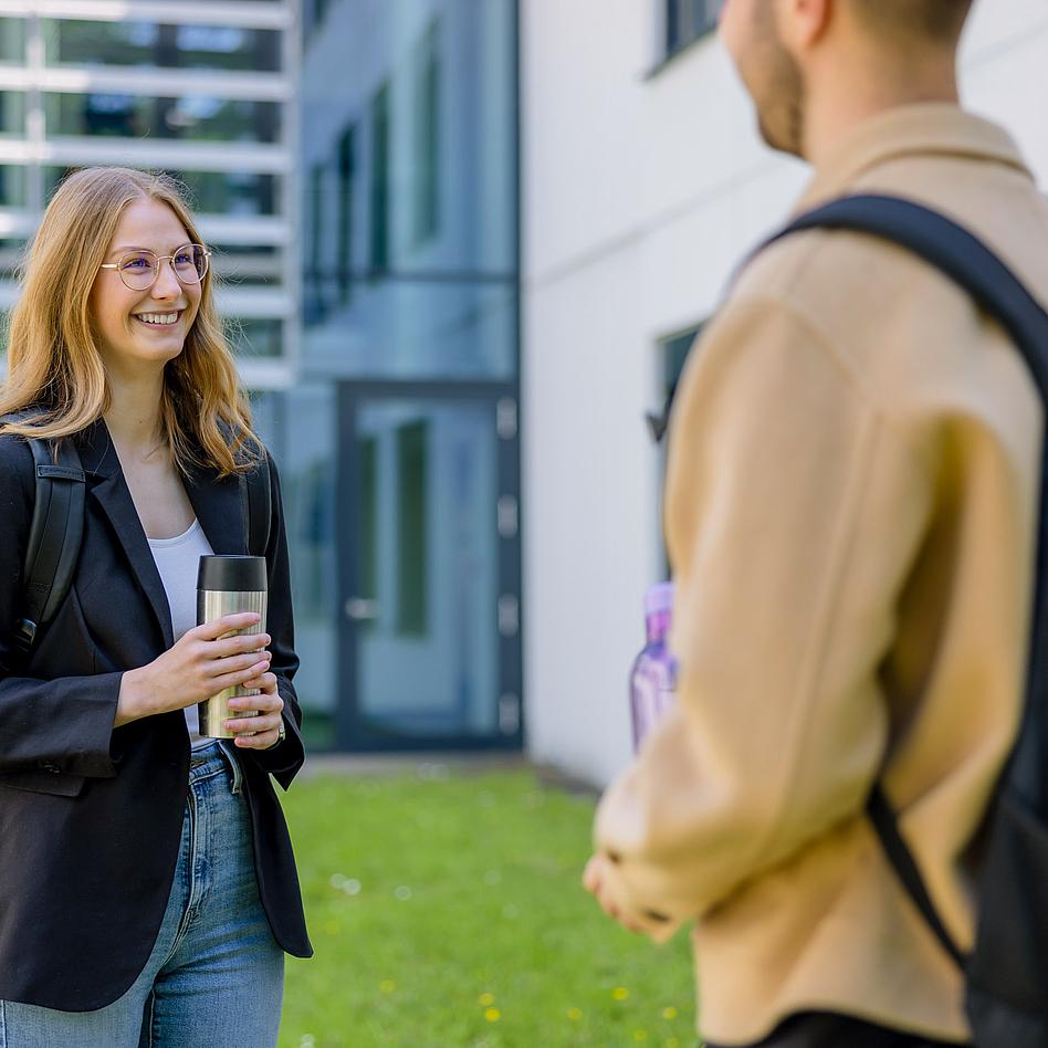 Eine junge Frau mit Brille und einem Kaffeebecher in der Hand steht einer Person gegenüber.