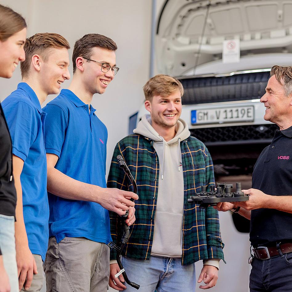 VOSS Mitarbeiter im Gespräch mit vier jungen Azubis, vor einem Pkw mit geöffneter Motorhaube. 