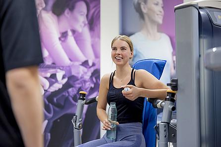 Junge Frau sitzt im VOSSletics, dem VOSS eigenen Fitnessstudio und unterhält sich mit ihrem Kollegen